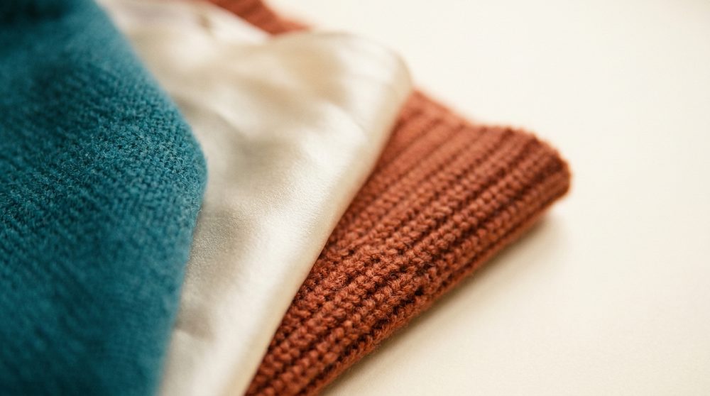 Three layers of cloth overlapping on a cream surface — teal wool, cream silk and an orange rib knit