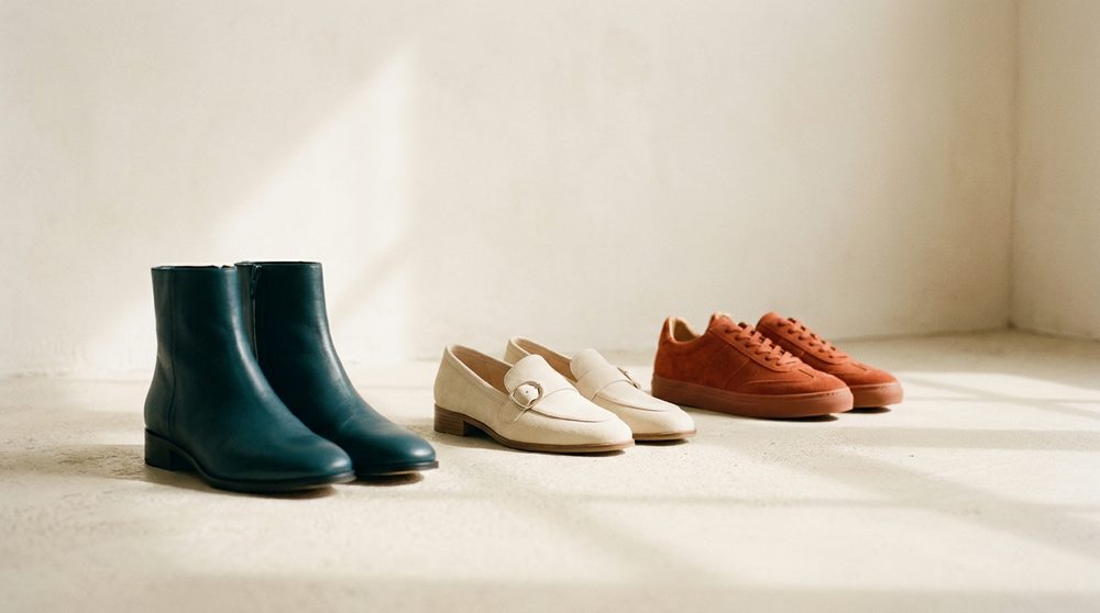 Three pairs of shoes in a row on a pale concrete floor: teal boots, cream loafers, orange trainers